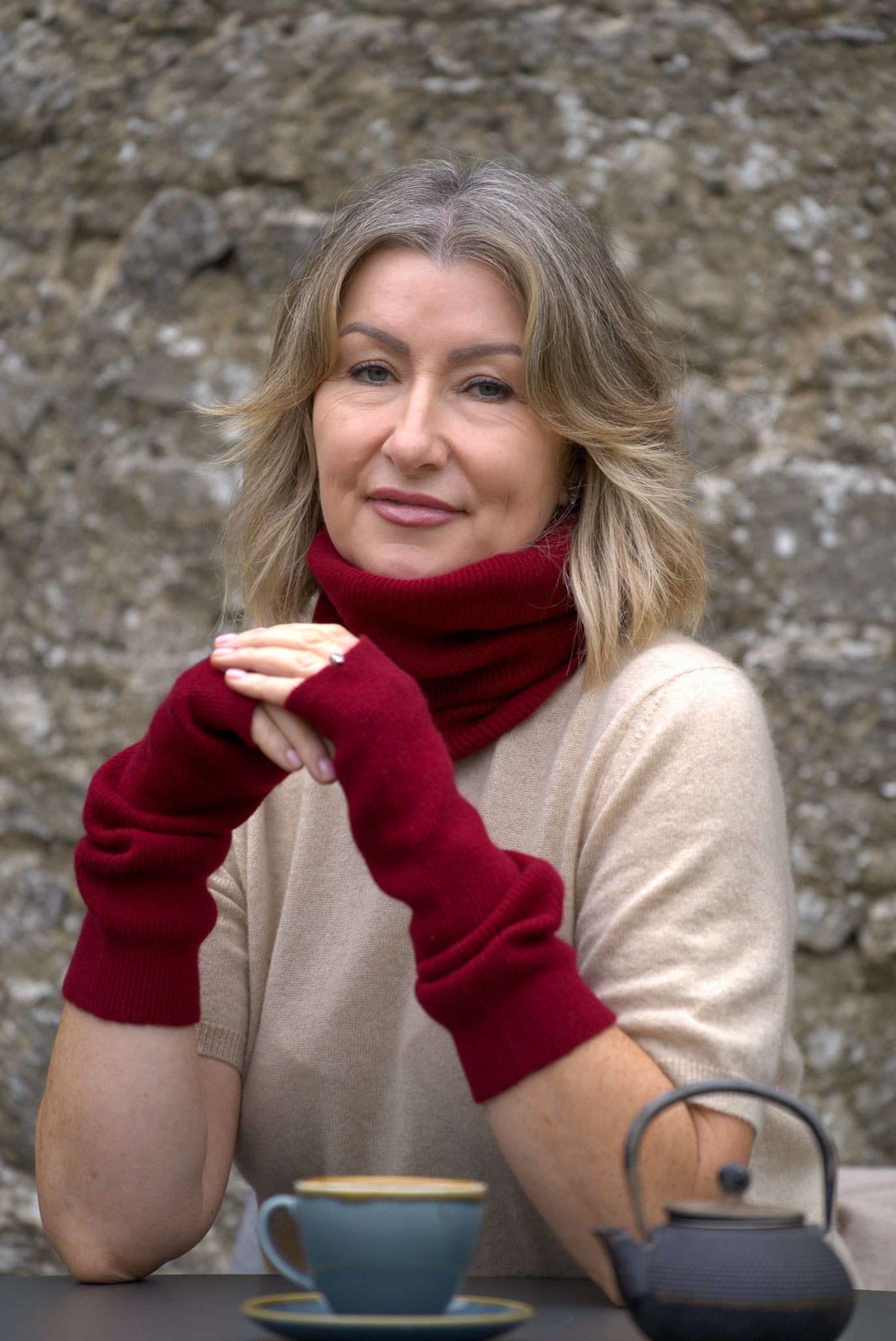 Woman wearing red arm warmers sitting at a table with a cup and teapot against a stone wall.